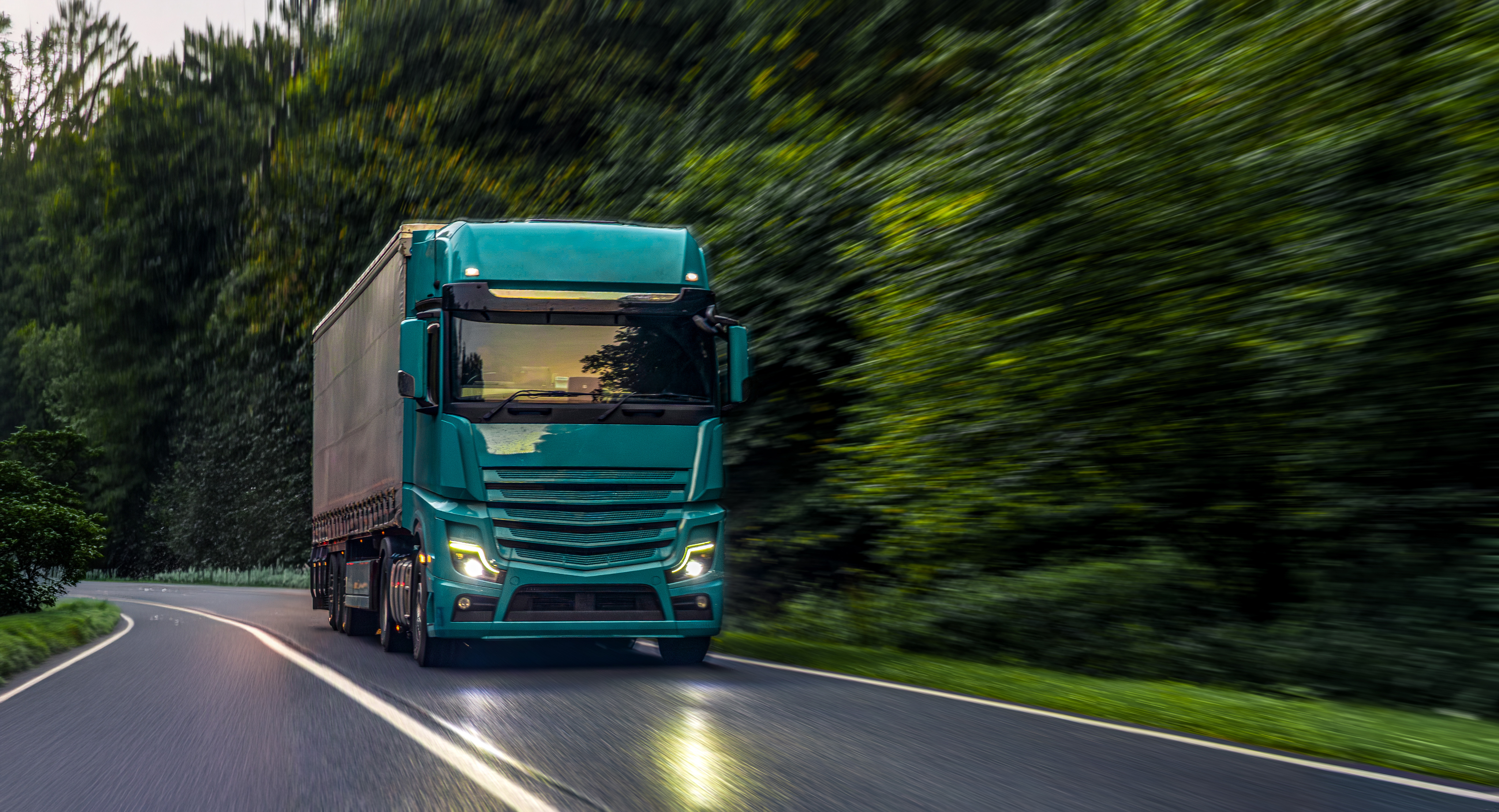 An articulated lorry travelling along a woodland road towards the camera. It is evening and the artic's head lights are lit